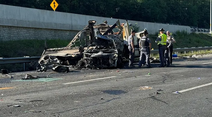 Emergency responders inspect a severely burned vehicle wreck on a highway, with debris scattered around and a guardrail in the background.