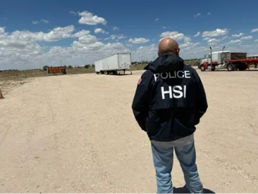 HSI police officer standing with back to camera on a dirt road, trucks and a desert landscape in the background.