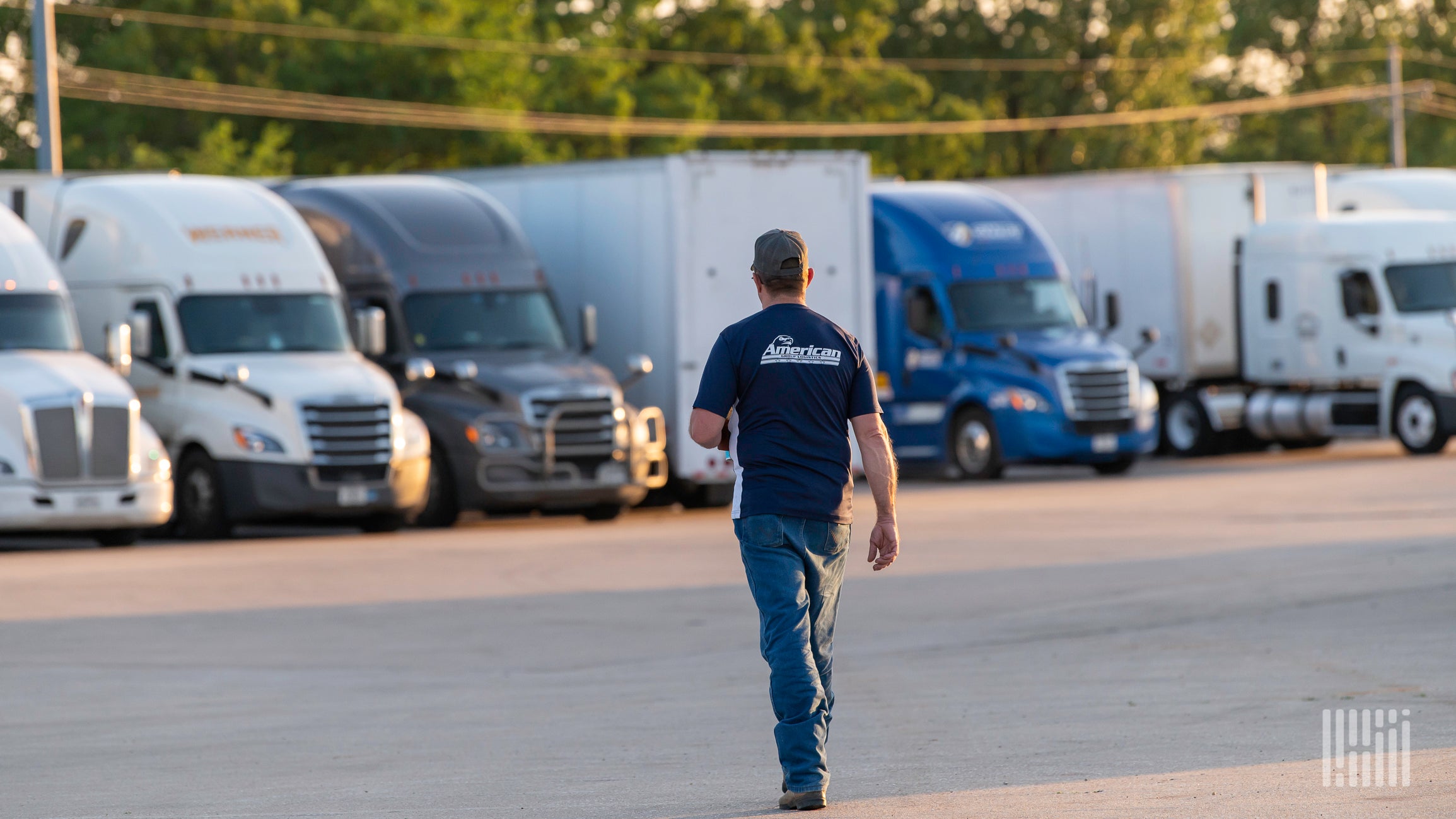 Man wearing a navy shirt and cap walks away between row of white, gray, and blue semi-trucks in a truck yard.