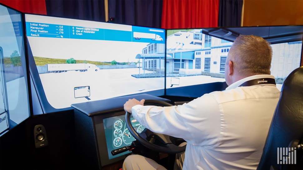 Trainee operating a driving simulator with curved screens showing a city street and vehicle dashboard.