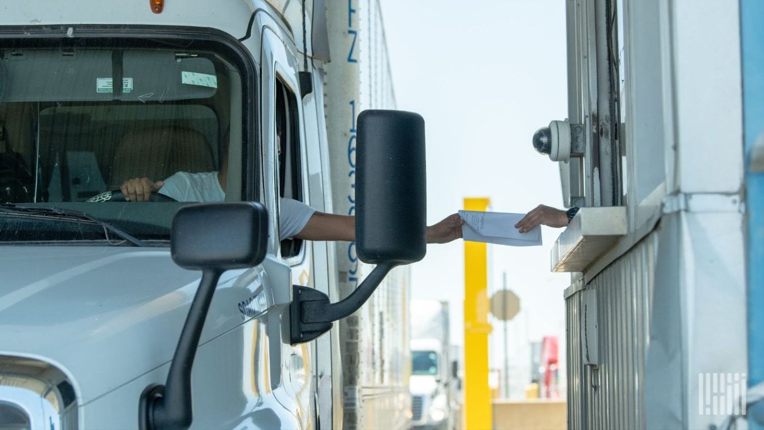Truck driver in a white cab hands an envelope to a toll booth attendant through a window.