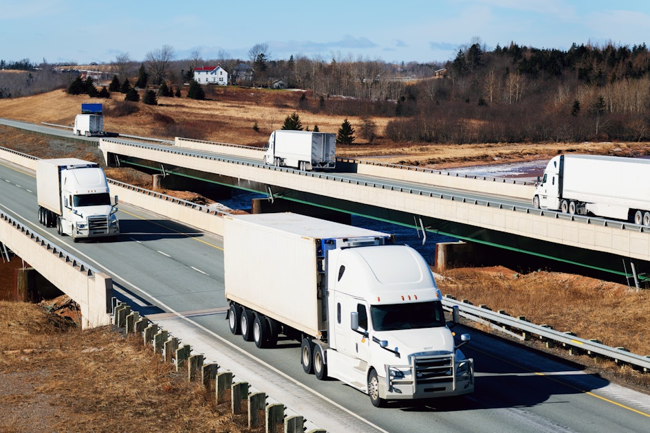 White semi-trucks driving on a multi-level highway through a rural landscape with a grassy hill and bare trees in the background.
