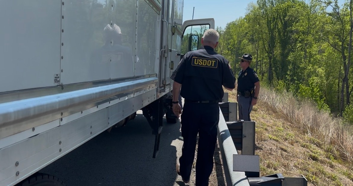 Two USDOT officers inspect a large white truck parked on the highway shoulder beside a metal guardrail.