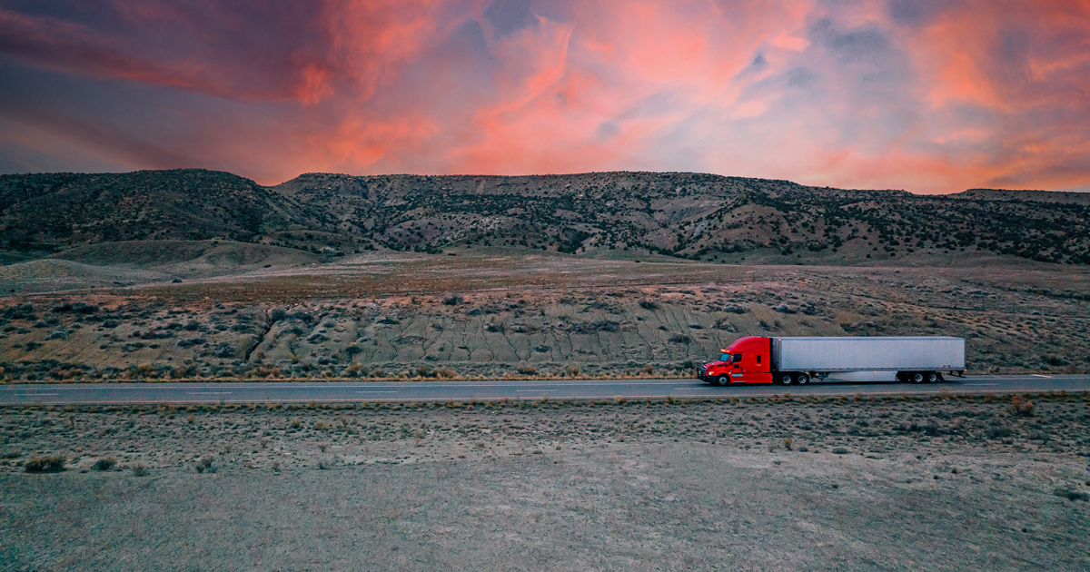 Red semi-truck with a white trailer driving on a two-lane highway through a desert landscape at sunset, with rocky hills in the background.