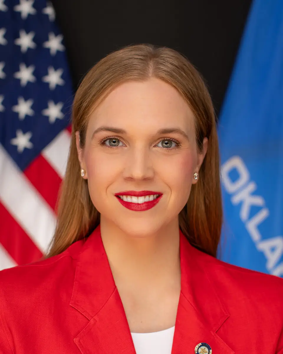 Head-and-shoulders portrait of a woman in a red blazer, smiling with red lipstick, with the American flag in the background.