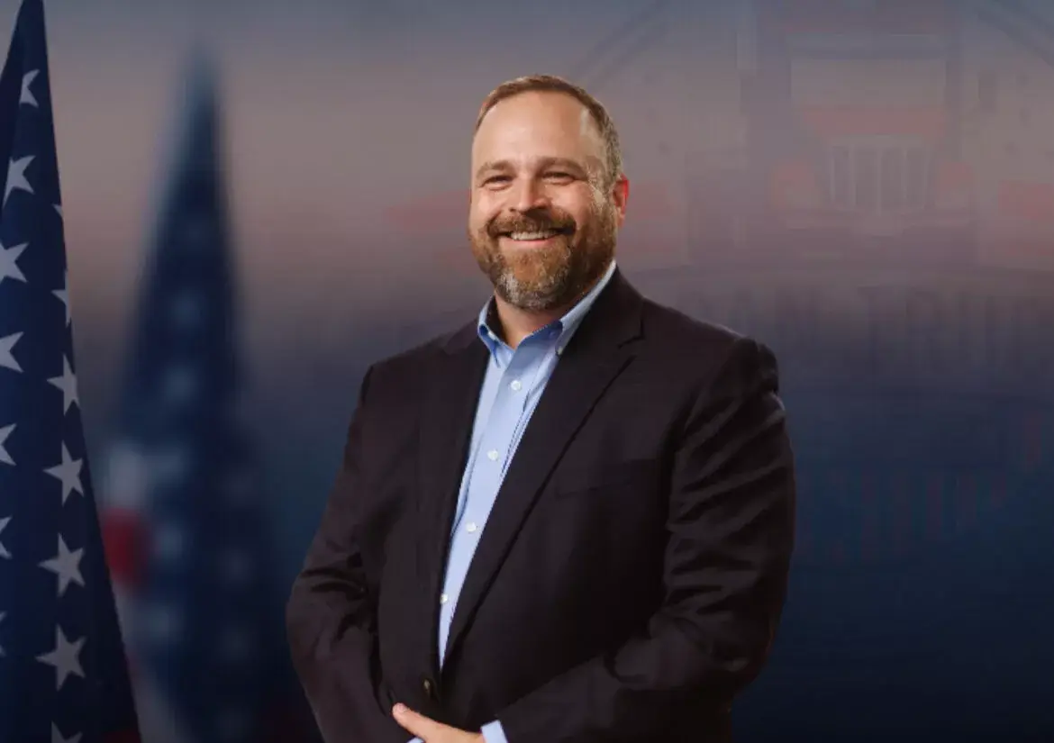 Smiling man in a dark suit and light blue shirt stands with hands folded in front, with the US flag and government backdrop behind him.