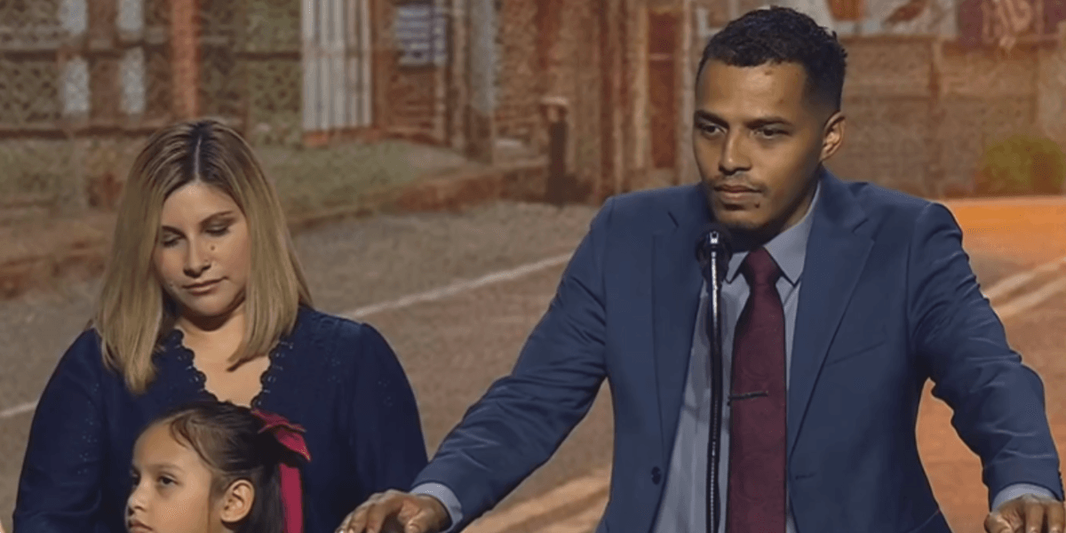 Man in a blue suit speaks into a microphone at a podium, with a woman and a young girl beside him on an outdoor street set.
