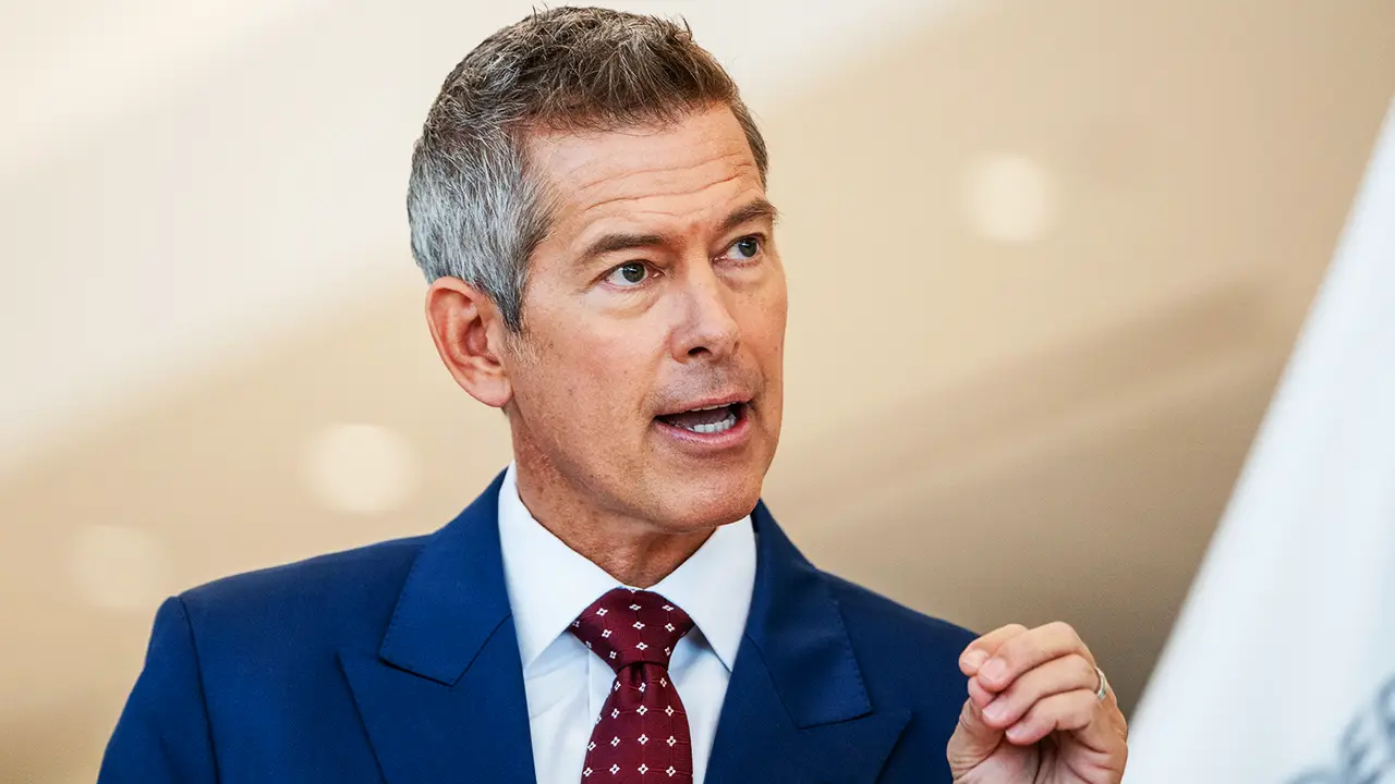 Middle-aged man in a blue suit and red patterned tie speaks, gesturing with his hand during a presentation/meeting.