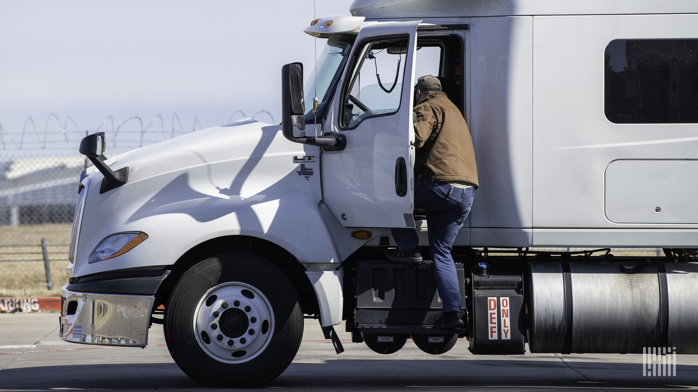 White semi-truck with driver climbing into the cab on a sunny day near a fence.