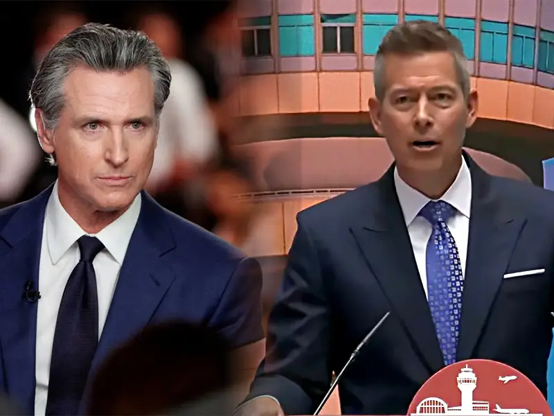 Two suited men on a TV set; one seated on the left, the other standing at a podium and speaking on the right.