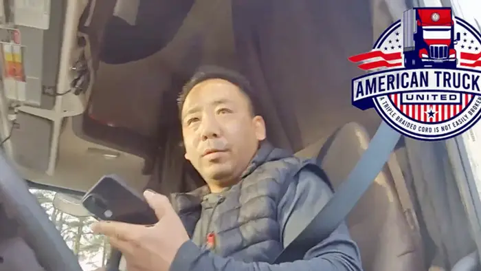 Truck driver in the cab wearing a seatbelt, looking at a smartphone, with an American Truck United logo in the top-right corner.