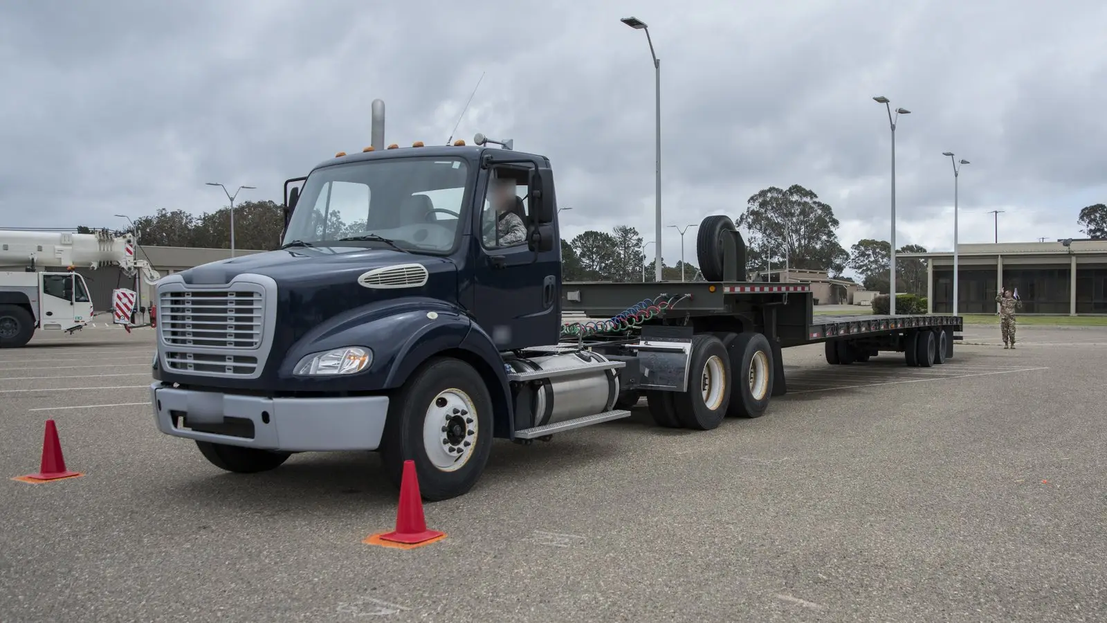 Dark blue semi-truck cab pulling an empty flatbed trailer past orange traffic cones on a paved training lot