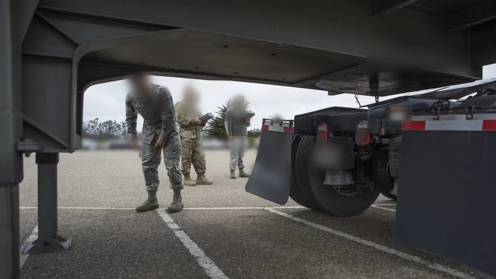 View from under a semi-trailer as uniformed airmen inspect a tractor-trailer on a paved CDL training lot