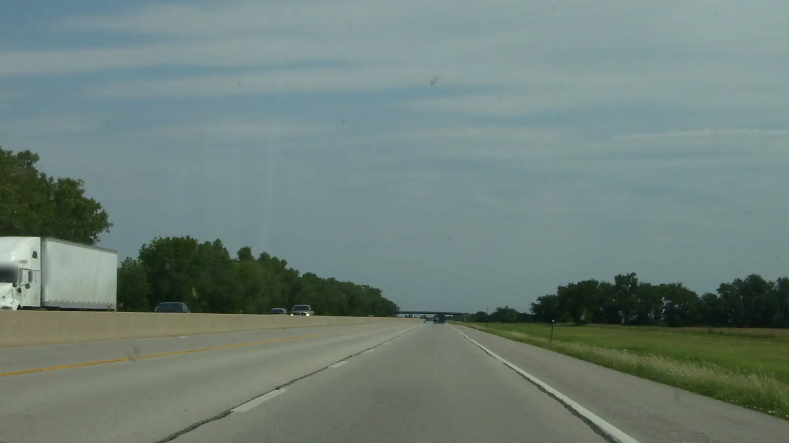 A Kansas interstate under a cloudy sky, with a white semi truck in the oncoming lanes beside green fields
