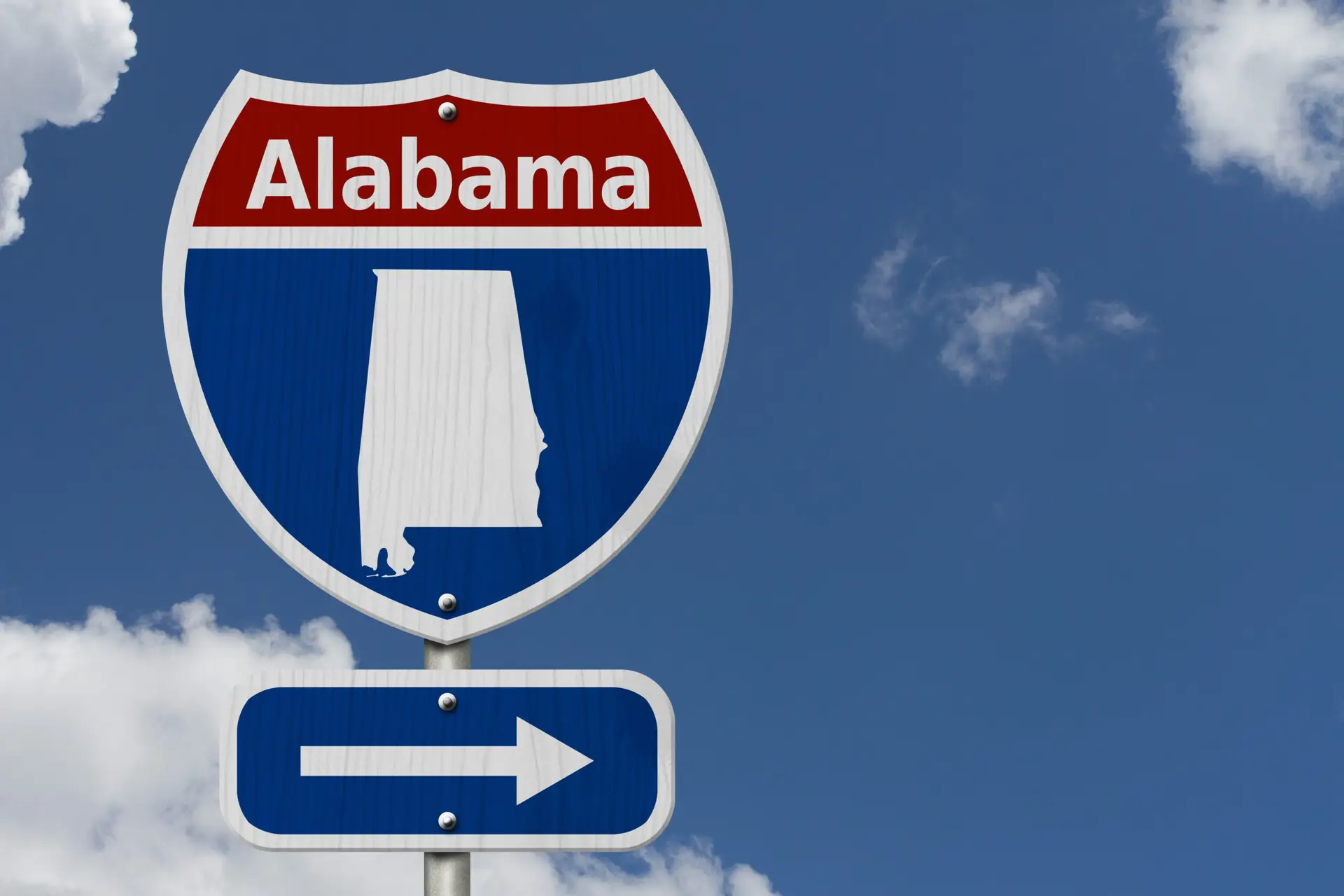 Red, white and blue Alabama interstate-style road sign with a white state outline, above a blue sign with a right-pointing arrow, against a blue sky with white clouds